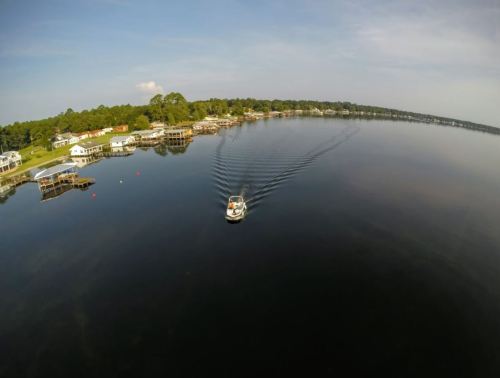 Aerial view of a calm lake with a boat leaving ripples, surrounded by houses and trees along the shoreline.