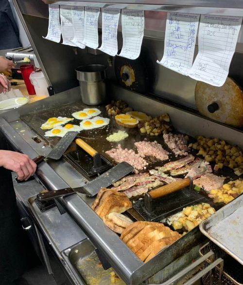 A busy kitchen grill with cooking eggs, meats, potatoes, and toast, alongside order tickets hanging above.