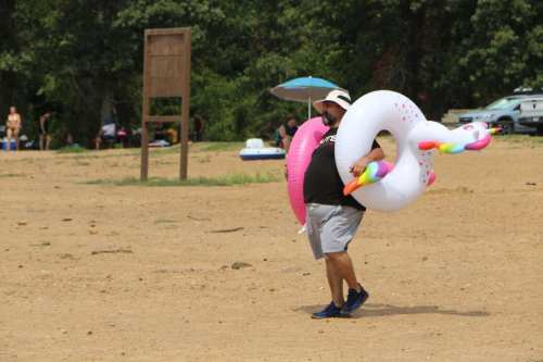 A man in a hat carries a large inflatable unicorn float at a sandy beach.