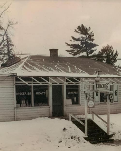 A vintage store with a sign reading "Lunches," surrounded by snow and trees, featuring windows displaying groceries and meats.