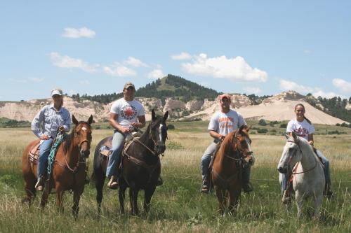 Four riders on horseback in a grassy field, with hills and blue sky in the background.