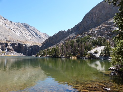 A serene mountain lake surrounded by rocky cliffs and lush green trees under a clear blue sky.