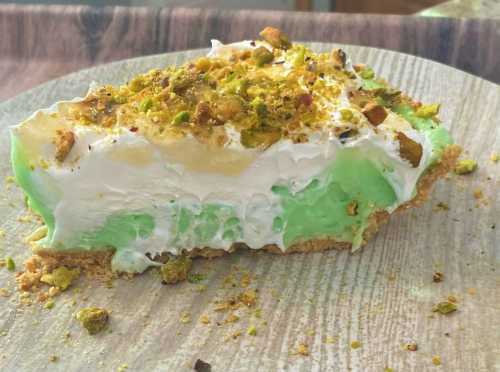 A slice of green pistachio pie topped with whipped cream and crushed pistachios on a wooden plate.