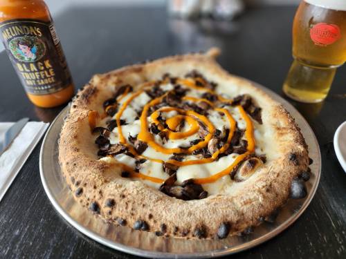 A pizza with a golden crust topped with mushrooms and a swirl of orange sauce, alongside a beer and hot sauce.