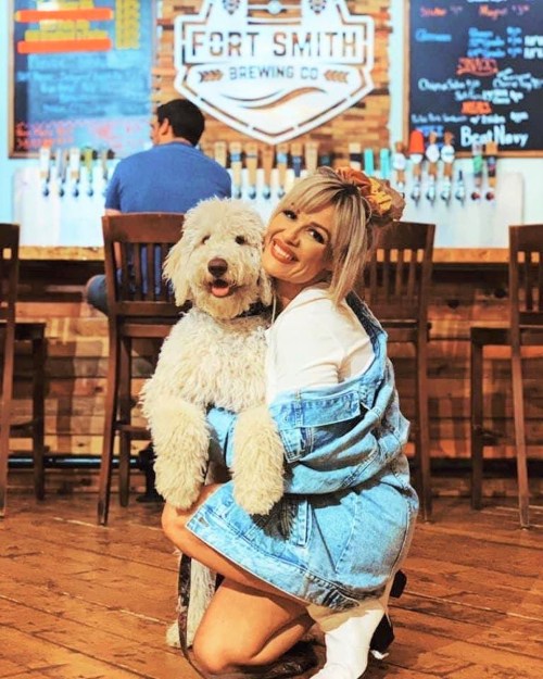 A woman in a denim jacket hugs a fluffy dog in a brewery with a rustic wooden interior.