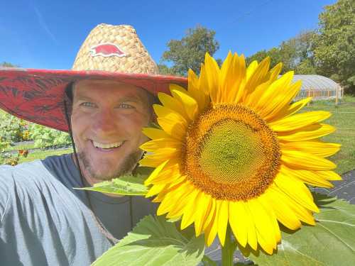 A smiling person in a straw hat poses next to a large sunflower in a sunny garden.