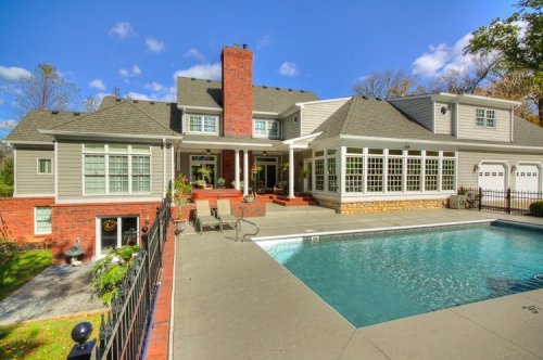 A spacious house with a pool, featuring a brick chimney and large windows, surrounded by greenery and a clear blue sky.