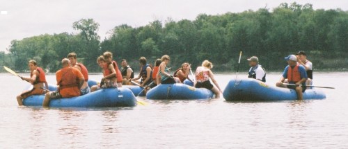 A group of people in life jackets paddling on blue rafts in a calm river surrounded by greenery.