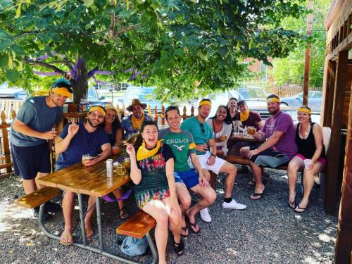 A group of friends wearing yellow bandanas, smiling and enjoying drinks at an outdoor table under a tree.