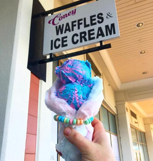 A hand holds a colorful ice cream cone in front of a sign that reads "Coney Waffles & Ice Cream."