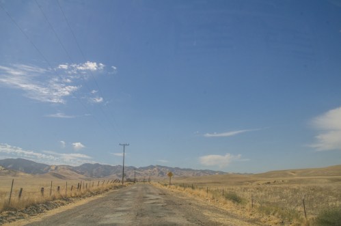 A rural road stretches into the distance, flanked by dry fields and mountains under a clear blue sky.
