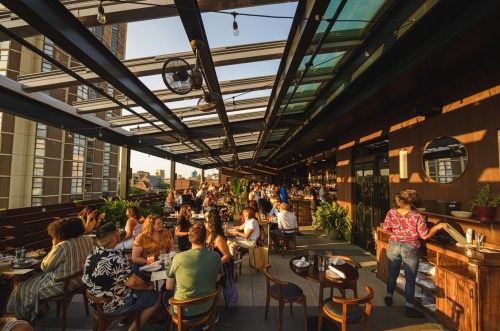 A bustling rooftop restaurant with diners enjoying meals under a glass canopy, surrounded by greenery and city views.