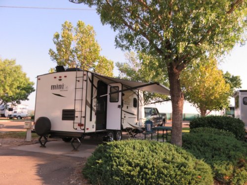 A small RV parked under trees at a campsite, with an awning extended and a table nearby.