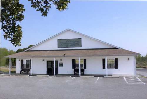 A white building with a sloped roof and green sign, featuring black shutters and a gravel parking lot.