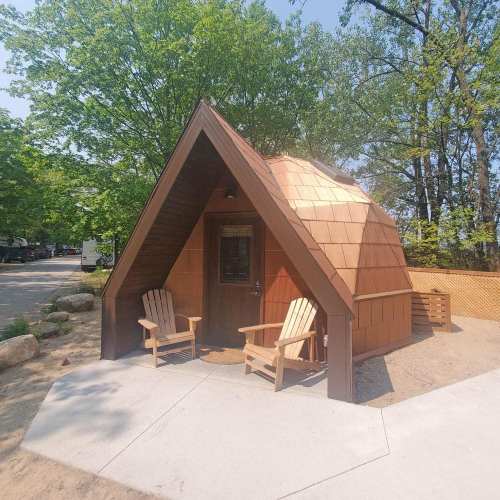 A small A-frame cabin with wooden shingles, surrounded by trees, featuring two outdoor chairs on a concrete patio.