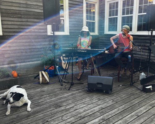Two musicians perform on a wooden deck, with a dog resting nearby and a rainbow effect in the background.