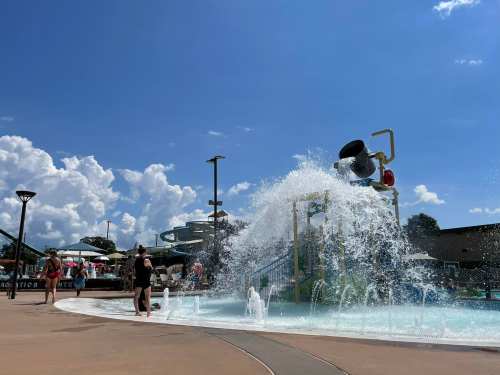 A water play area with a large splash feature, people enjoying the sun, and fluffy clouds in a blue sky.