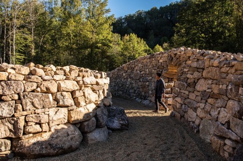 A person walks through a stone pathway surrounded by tall trees and sunlight filtering through the landscape.