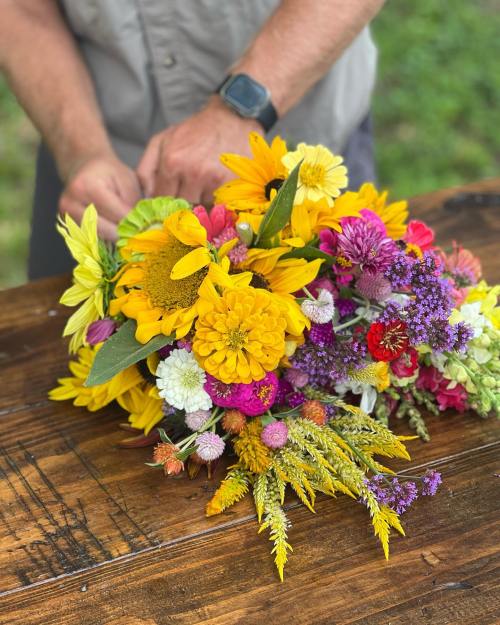 A person arranging a vibrant bouquet of sunflowers and colorful wildflowers on a wooden table.