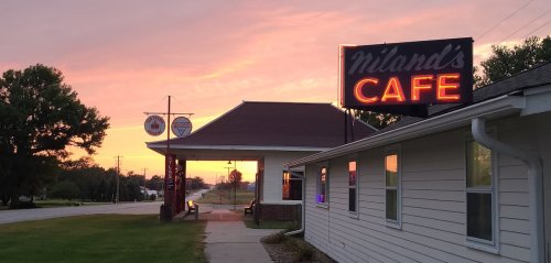 A sunset view of Miland's Cafe with a neon sign, surrounded by trees and a quiet road.