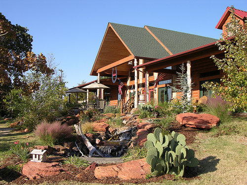 A charming log cabin with a green roof, surrounded by landscaped gardens and a small water feature.