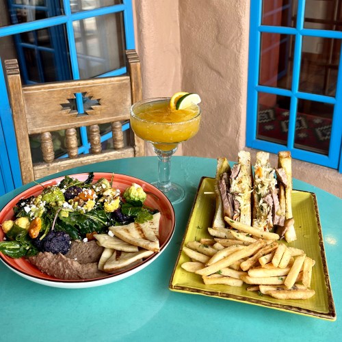 A colorful meal featuring a salad, sandwich, fries, and a margarita on a turquoise table with blue windows in the background.