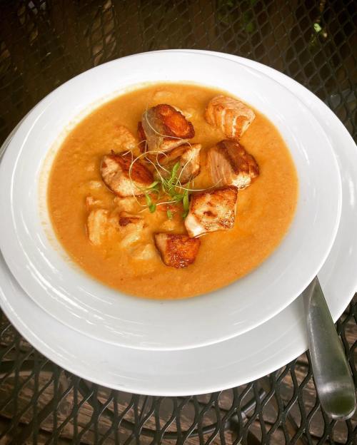 A bowl of creamy soup with chunks of grilled fish and garnished with microgreens, served on a white plate.