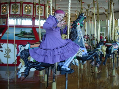 A child in a purple dress and pink hair joyfully rides a carousel horse, surrounded by colorful carousel decorations.
