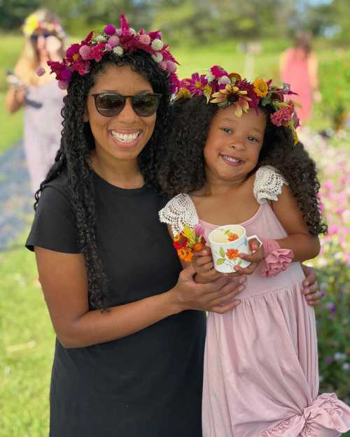 A smiling woman and girl wear flower crowns, holding a cup and flowers, surrounded by a colorful garden.