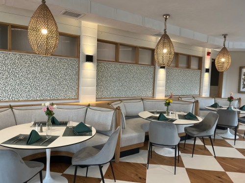Modern restaurant interior featuring round tables, gray chairs, and decorative pendant lights. Floral centerpieces on tables.