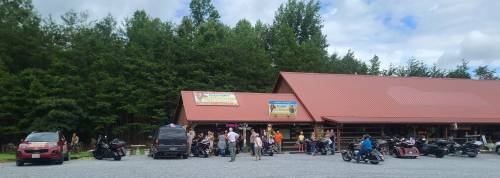 A group of people and motorcycles gathered outside a rustic building with a red roof, surrounded by trees.