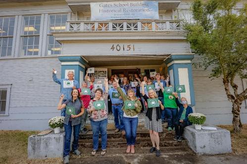 A group of people stands on the steps of a historic building, holding signs and smiling, celebrating a restoration project.