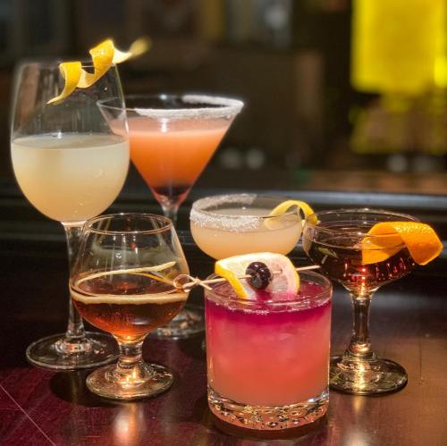 A variety of colorful cocktails in elegant glasses, garnished with fruit and citrus twists, arranged on a bar.