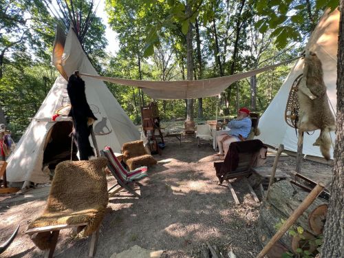 A campsite with two tipis, wooden chairs, and animal pelts hanging, surrounded by trees and a sunny outdoor setting.