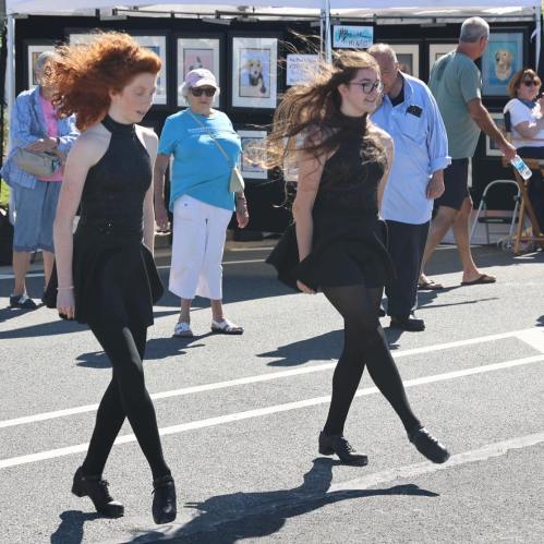 Two girls in black dresses and tights perform a dance routine at an outdoor event, with spectators in the background.