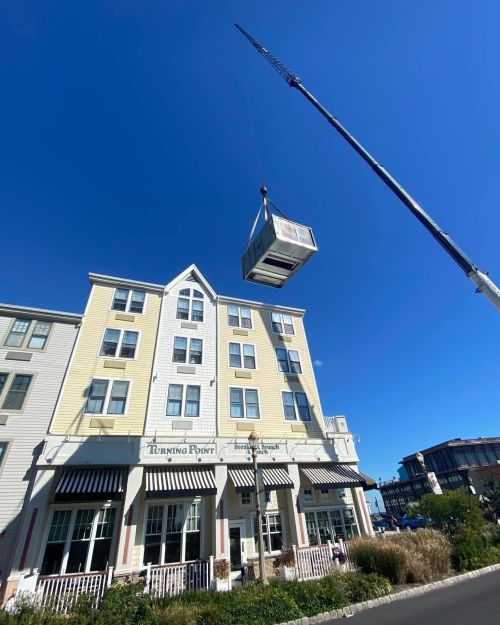 A crane lifts a large structure above a multi-story building against a clear blue sky.