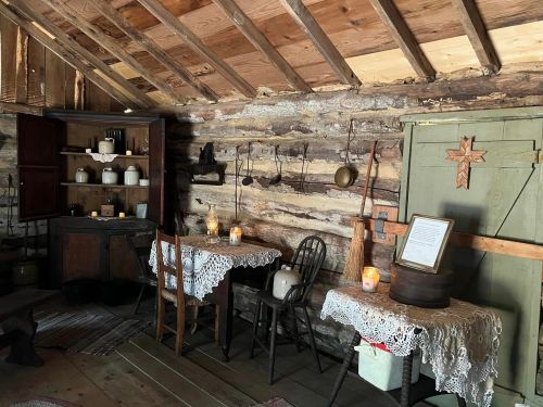 Cozy interior of a rustic cabin with wooden walls, tables with lace, candles, and vintage kitchenware.