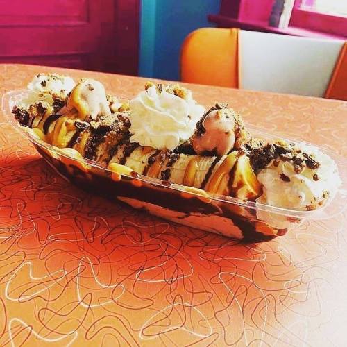 A colorful dessert bowl with bananas, whipped cream, chocolate sauce, and nuts on a vibrant orange table.