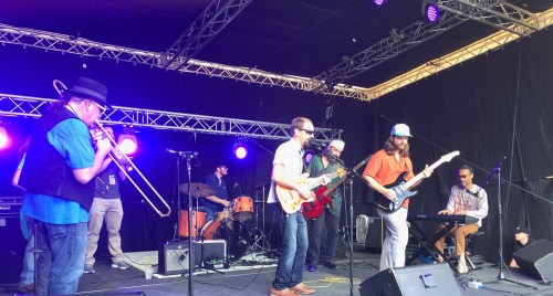 A lively band performs on stage with various instruments, including a trombone, guitar, and keyboard, under colorful lights.