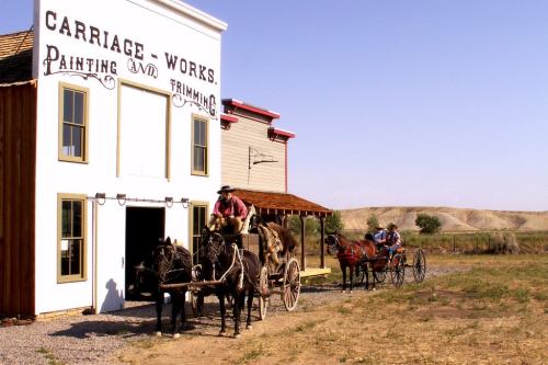 A historic carriage works building with two horse-drawn carriages outside, set against a clear blue sky and prairie landscape.