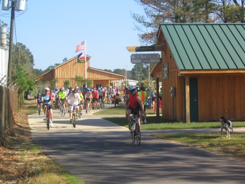 A group of cyclists riding past wooden buildings, with an American flag flying and a dog on the path.