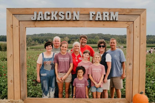 A group of people poses together in front of a wooden frame labeled "JACKSON FARM" with a scenic farm background.