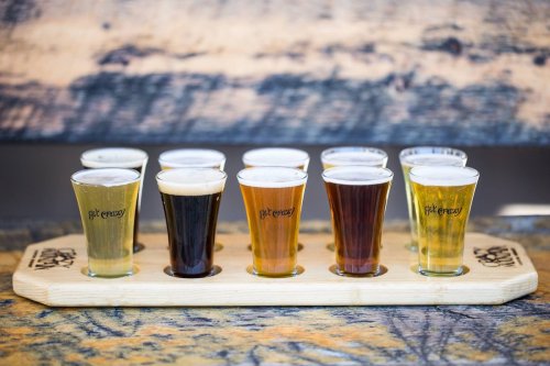 A wooden flight board holds ten small glasses of various craft beers, showcasing different colors and styles.