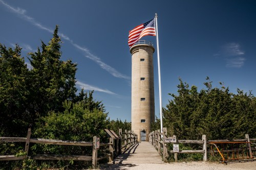 A tall lighthouse with an American flag, surrounded by greenery and a wooden fence under a clear blue sky.