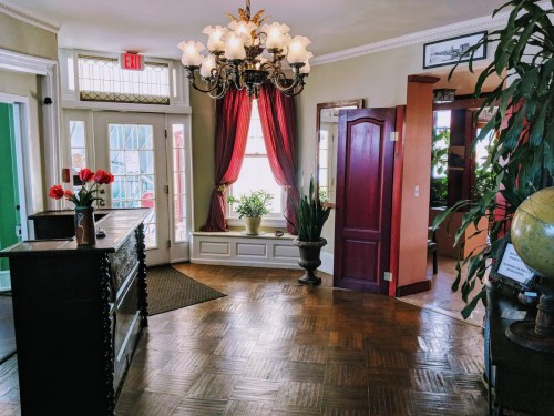 A cozy entryway with a chandelier, red curtains, plants, and a reception desk, featuring warm wooden flooring.