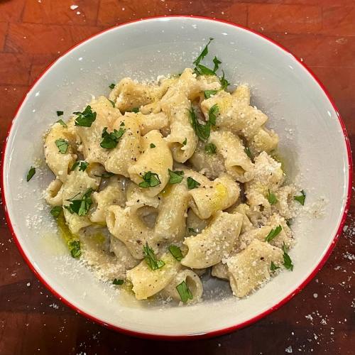 A bowl of pasta topped with herbs, grated cheese, and a drizzle of olive oil on a wooden surface.