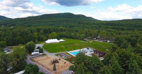 Aerial view of a green landscape featuring a playground, pool, and a large white tent surrounded by trees and mountains.