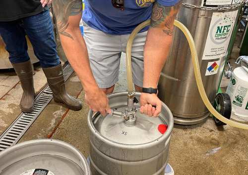 A person is connecting a hose to a keg, with another person in the background wearing rubber boots.