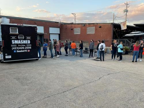 A food truck named "Smashed" with a line of people waiting outside in an urban setting during sunset.
