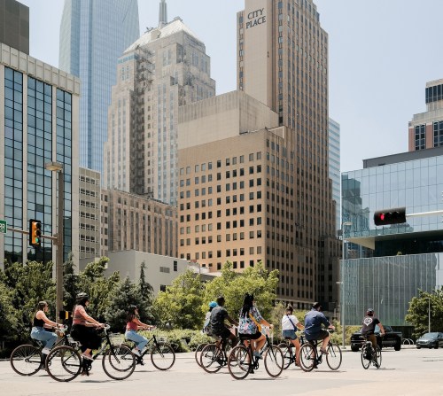 A group of cyclists riding through a city intersection with tall buildings in the background on a sunny day.